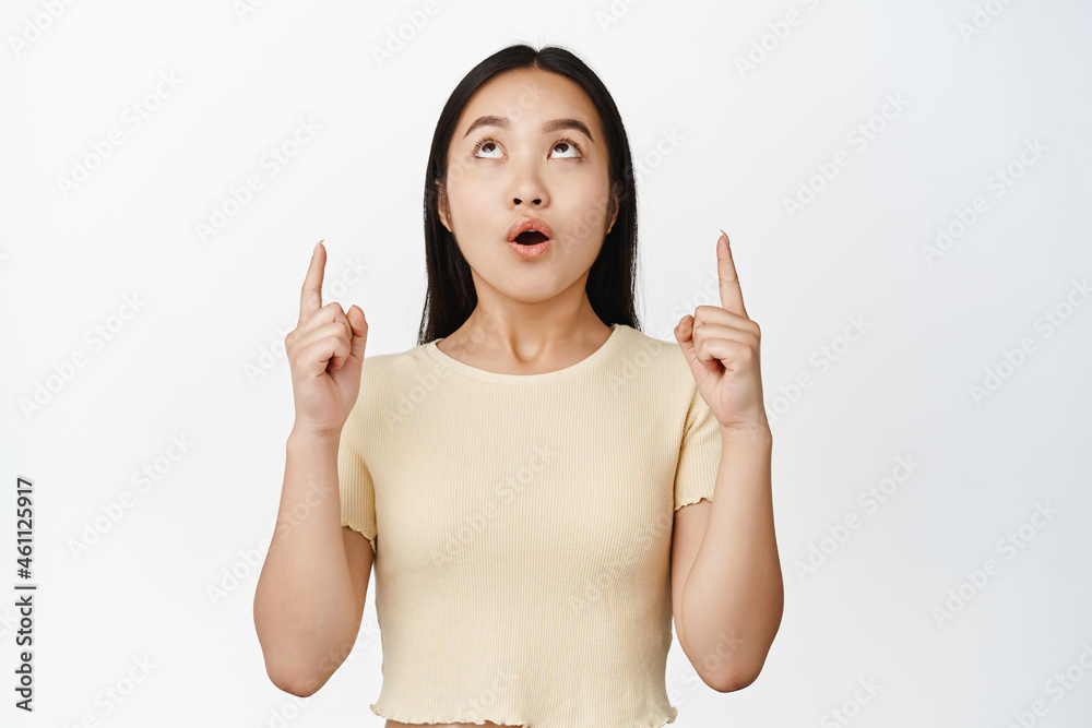 Amazed asian girl pointing fingers up, looking fascinated at price discount, showing loog on top, standing over white background