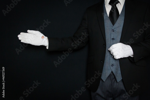 Fotografi Portrait of Butler in Dark Suit and White Gloves Standing with Welcoming Gesture on Black Background