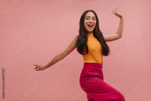 Cheerful asian girl with loose dark hair having fun and posing on pink background. High quality photo of young beauty dancing in studio, in summer colorful clothes.