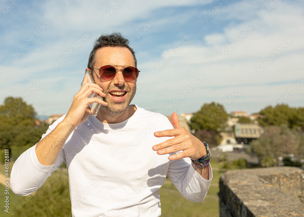 white man with sunglasses and a white T-shirt talks on the phone in the city with a exited expression