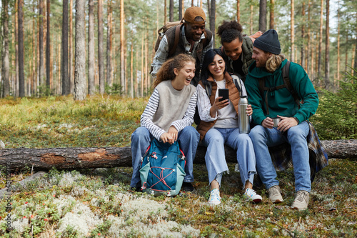 Full length portrait of diverse group of friends sitting on log in forest and using smartphone while enjoying hiking in Autumn, copy space