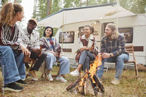 Low angle view at diverse group of modern young people enjoying camping outdoors with trailer van and sitting by fire, copy space