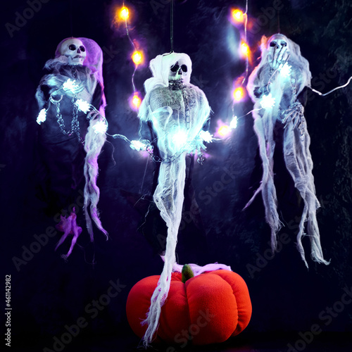 Halloween decoration three skeletons in a multicolored cape decorated with a garland of glowing pumpkins and spiders. There is a decorative pumpkin at the bottom