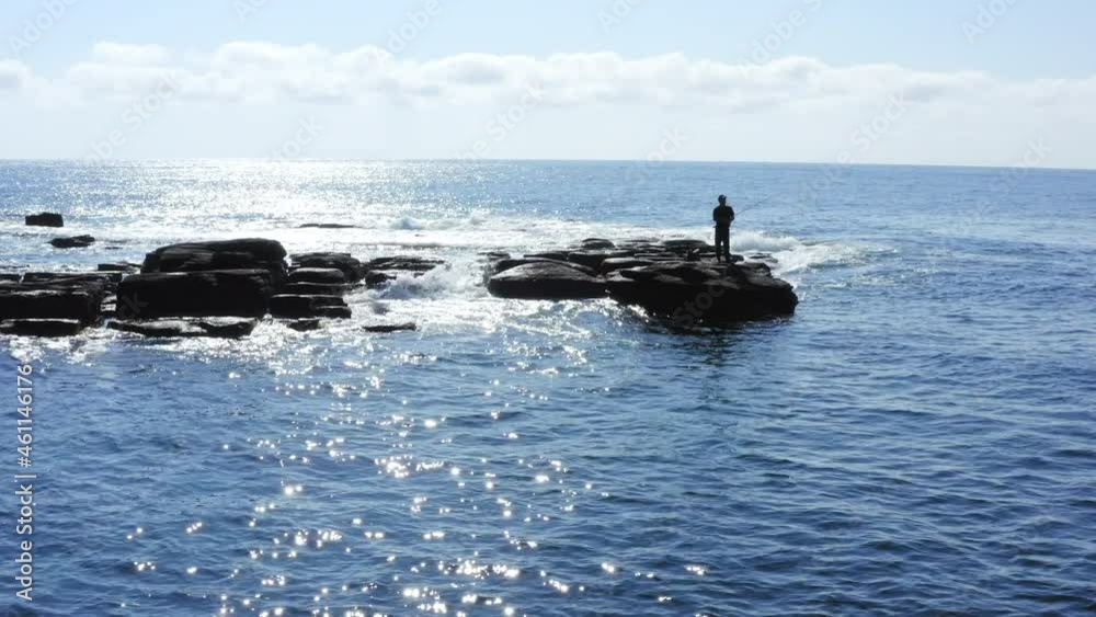 Aerial panoramic view of a man fishing at the seaside. High rocks and the amazing blue ocean are on the background, foamy waves are splashing at his feet. Male hobby Aerial panoramic view of a man fishing at the seaside. High rocks and the amazing blue ocean are on the background, foamy waves are splashing at his feet. Male hobby