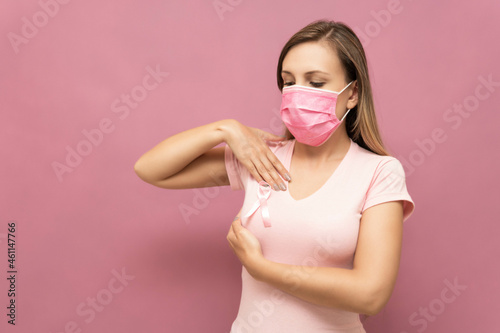 Young blonde woman with pink mask does her breast cancer self-exam wearing a pink t-shirt with the cancer ribbon icon.