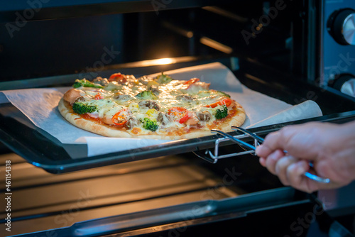 Cooking italian pizza in electric convection oven. Woman open the oven and Italian Pizza with mushrooms, tomato sauce, cheese, sausage, bacon, bell pepper, ham, pineapple, broccoli out from machine.