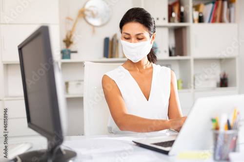 Young businesswoman in medical mask and white dress working with laptop and papers in office....