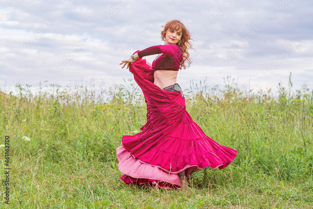 Young beautiful red-haired girl in a red ballroom dress is dancing an incendiary dance on a green meadow against a blue sky. Young beautiful red-haired girl in a red ballroom dress is dancing an incendiary dance on a green meadow against a blue sky.