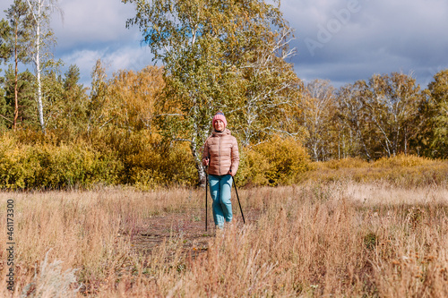 elderly sixty-year-old woman in sportswear is engaged in Nordic walking with sticks in the forest in the autumn © Alyona