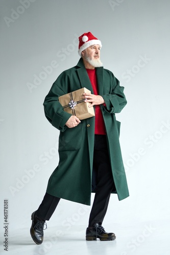 Full length shot of stylish mature man with Christmas hat wearing casual outfit looking away, holding gift box while posing isolated over light gray background