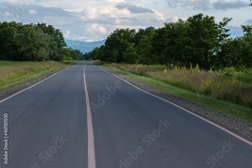 A suburban asphalt road with three solid lines of pink road markings. In the background of the mountain and the blue sky with clouds. The concept of travel.