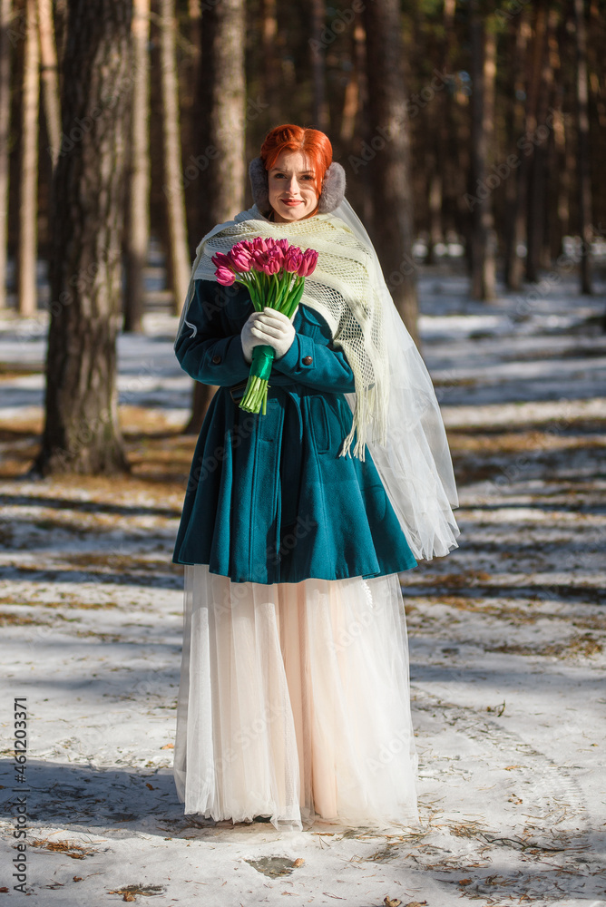 beautiful girl in a green coat and a wedding dress with a bouquet of flowers in the forest in winter