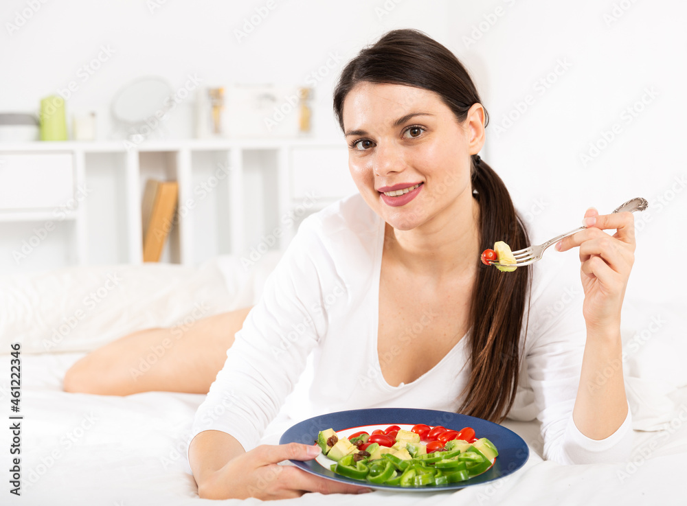Portrait of brunette lady in white dress eating vegetable salad in bed at home alone