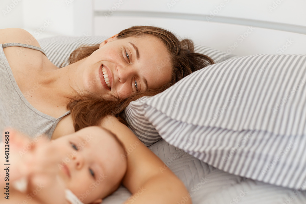 Indoor shot of female lying in bed with infant baby, expressing positive emotions and happiness, mother looking at her daughter or son with charming smile.