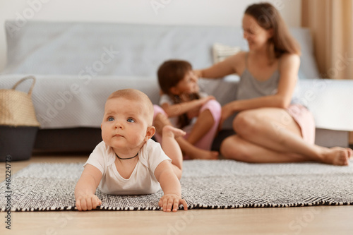 Indoor shot of cute adorable toddler girl lying on her belly on floor on carpet, looking up with interest, having curious facial expression, mother and sister on background sitting near sofa.