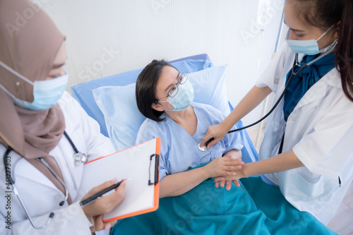 Portrait of two beautiful and cheerful female doctors wearing labcoat and stethoscope embracing senior patient sitting on hospital bed looking at camera