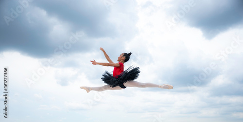 Cute little girl practicing ballet dance form wearing uniform and wooden shoes with a pose in the air isolated against a cloudy sky during day