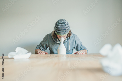 Ill man with long hair and hand made wool hat using a steam inhaler on wooden table during flu season
