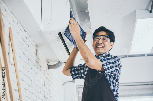 Young Asian male technician wearing safety glasses and apron cleaning air conditioner by removing the filter and fixing it back for further use