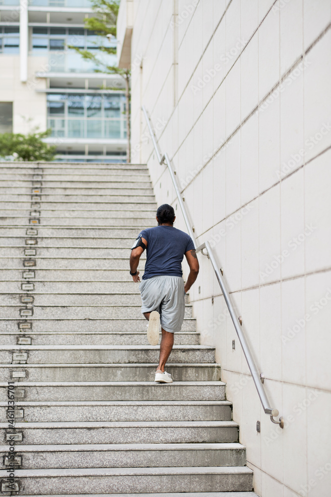 Rear view of sporty man with phone arm band running up stairs while practicing stair workout in city