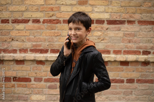 Photo of teenage boy aged 11-12 is talking on smartphone against background of brick wall in city, smile on his face