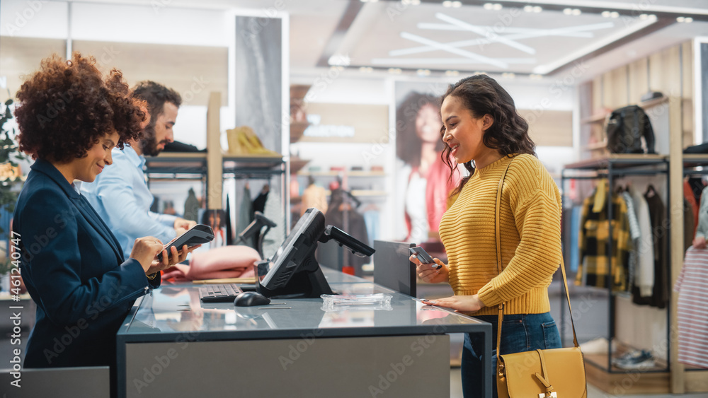 Clothing Store Checkout Cashier Counter: Woman and Male Retail Sales Managers Accept NFC Credit Card Payment from a Young Stylish Female Customer for New Stylish Clothes. - obrazy, fototapety, plakaty 