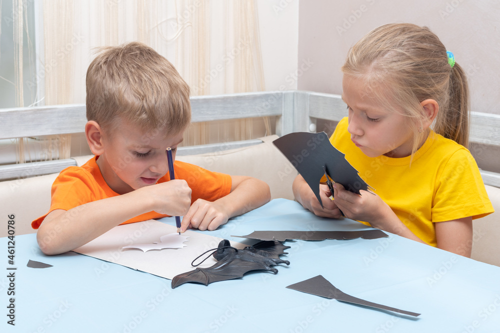 Boy and girl cut Halloween bats out of black paper with scissors at home. Children make bats out of black paper Boy and girl cut Halloween bats out of black paper with scissors at home. Children make bats out of black paper