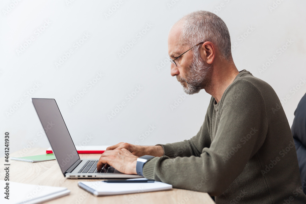 Adult gray-haired man working on a laptop at a desk, photo in profile Adult gray-haired man working on a laptop at a desk, photo in profile