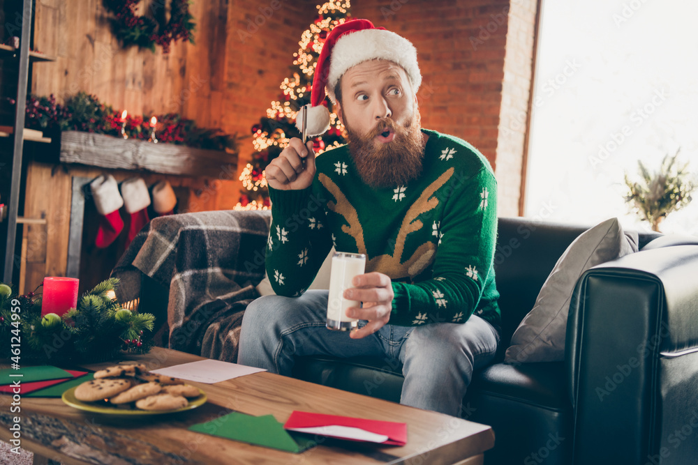 Photo of excited genius bearded guy write letter drink milk have idea wear santa hat sweater decorated home indoors