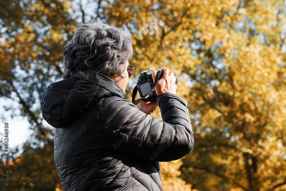 Back view of senior woman photographer using digital camera photographing autumn landscape in nature outdoors. Elderly people and technology, retirement hobby concept. Selective focus on retired woman