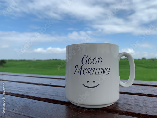 Morning greeting with a positive happy smile on white coffee or tea cup. Morning coffee concept  with white mug on the wooden table with bright summer blue sky and green field background. Good morning photo