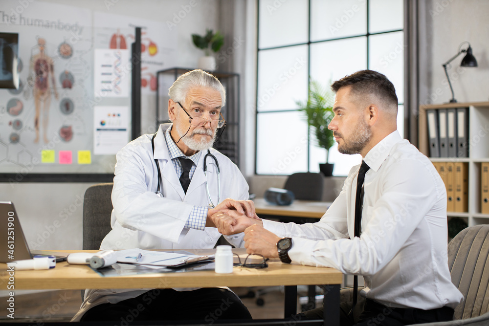 Qualified senior doctor measuring pulse on patient wrist while sitting together at desk. Caucasian businessman getting medical examination during visit to therapist. Concept of regular check up. Qualified senior doctor measuring pulse on patient wrist while sitting together at desk. Caucasian businessman getting medical examination during visit to therapist. Concept of regular check up.