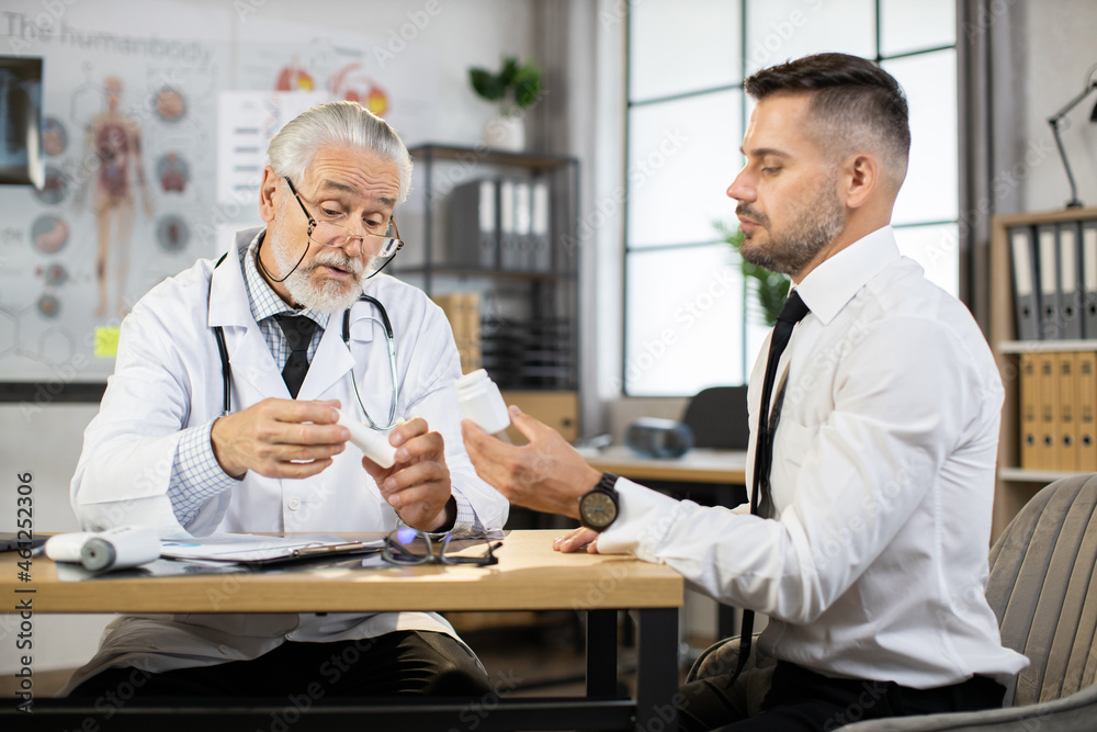 Mature therapist in white lab coat and eyewear consulting male patient in formal clothes about pills. Doctor prescribing medicine while sitting at desk with caucasian businessman.