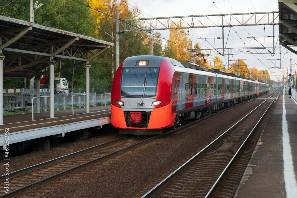 Naklejka premium Front view of modern russian intercity high speed passenger train on railroad at sunset, autumn forest and station platform on background. Commercial suburban railroad transportation concept