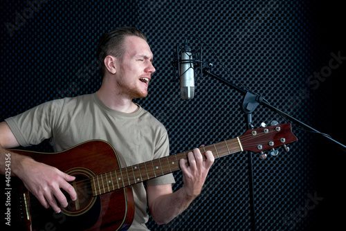 Young caucasian man sings while playing an acoustic guitar in front of black soundproofing walls. Musicians producing music in professional recording studio.