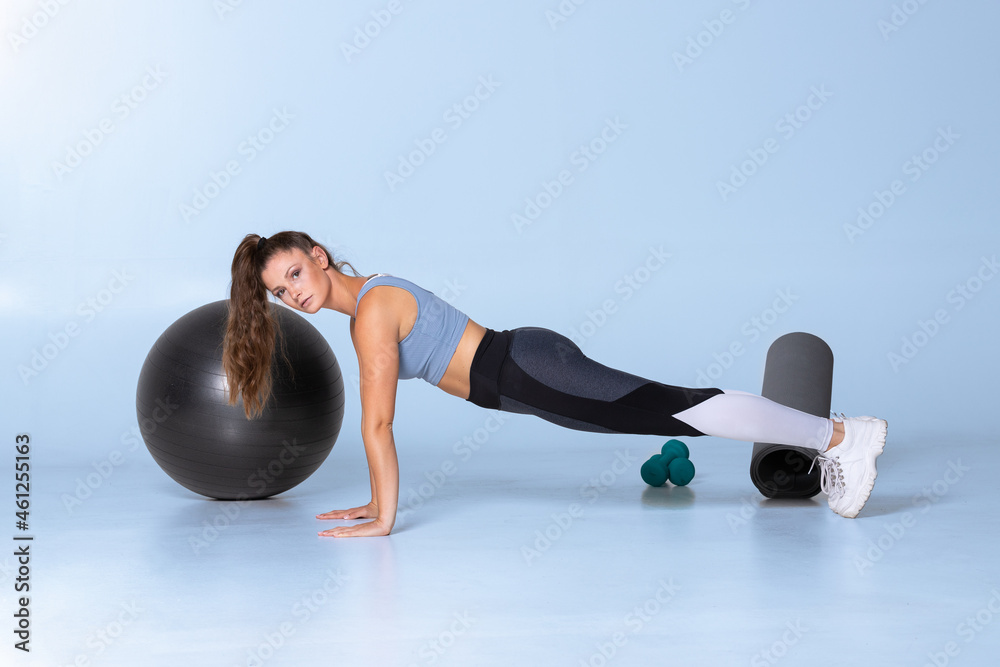 female fitness trainer standing in plank with strong back on blue background showing exercise looking at camera. Woman core exercise in gym with pony tale ball mat and dumbbells
