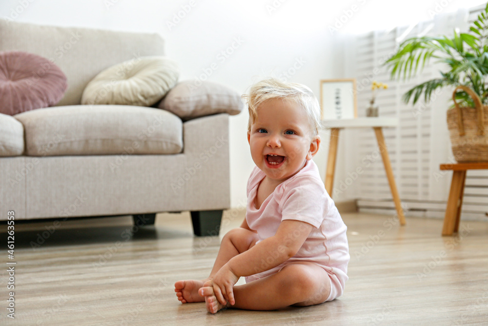 Portrait of smiling one year old child sitting on the wooden floor of living room. Interior background. Adorable blonde little girl in pink crawlers at home. Close up, copy space for text. Portrait of smiling one year old child sitting on the wooden floor of living room. Interior background. Adorable blonde little girl in pink crawlers at home. Close up, copy space for text.