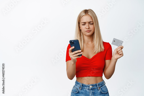 Sad blond teen girl holds mobile phone and credit card, having trouble with online purchase, standing over white background