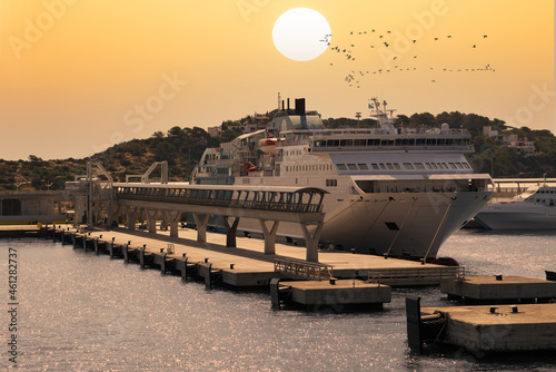 A ferry is moored at the terminal in the Spanish port of Ibiza. It is a summer morning and behind the ferry the morning sun is in the orange sky. A flock of birds flies by.
