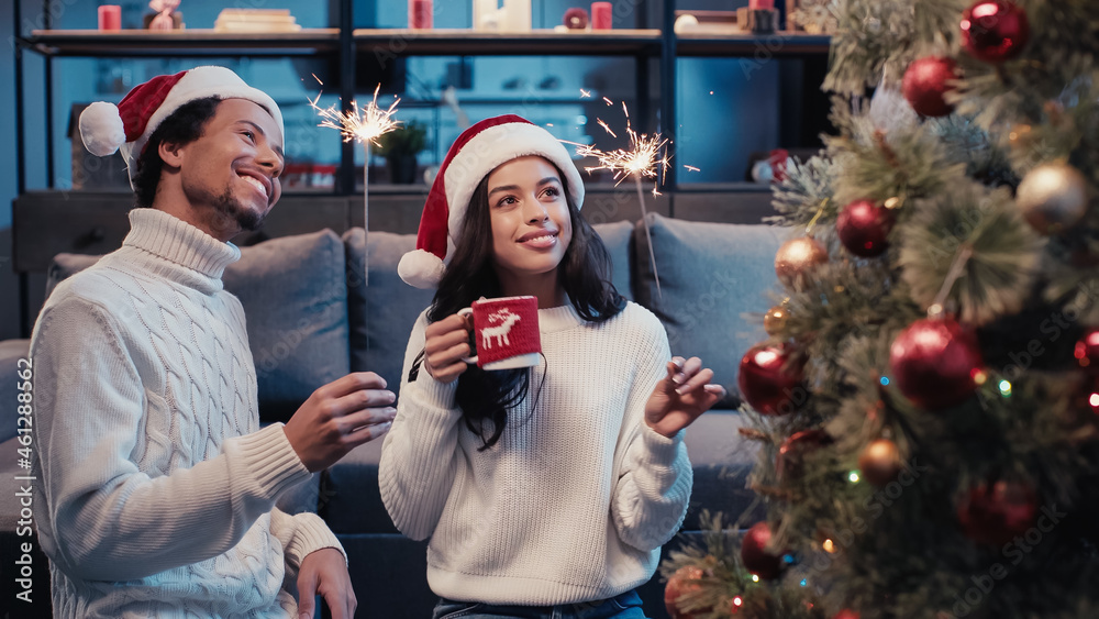 joyful african american couple in santa hats holding cup and sparklers while looking at blurred christmas tree