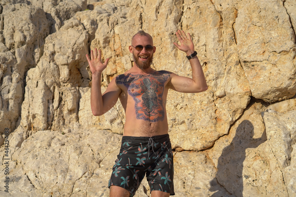 Young tattooed sporty man on the beach on a hot summer sunny day against the backdrop of a mountain posing in sunglasses