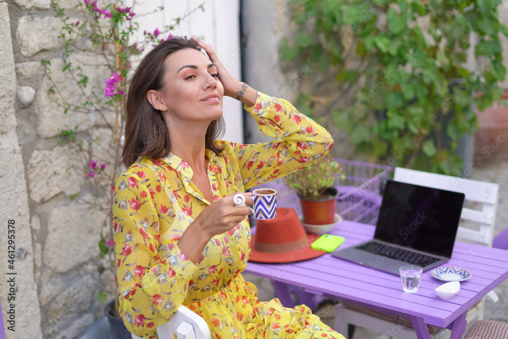 Fototapeta premium Young woman in a dress in a bright street cafe with a laptop drinks Turkish coffee, works remotely on her own schedule