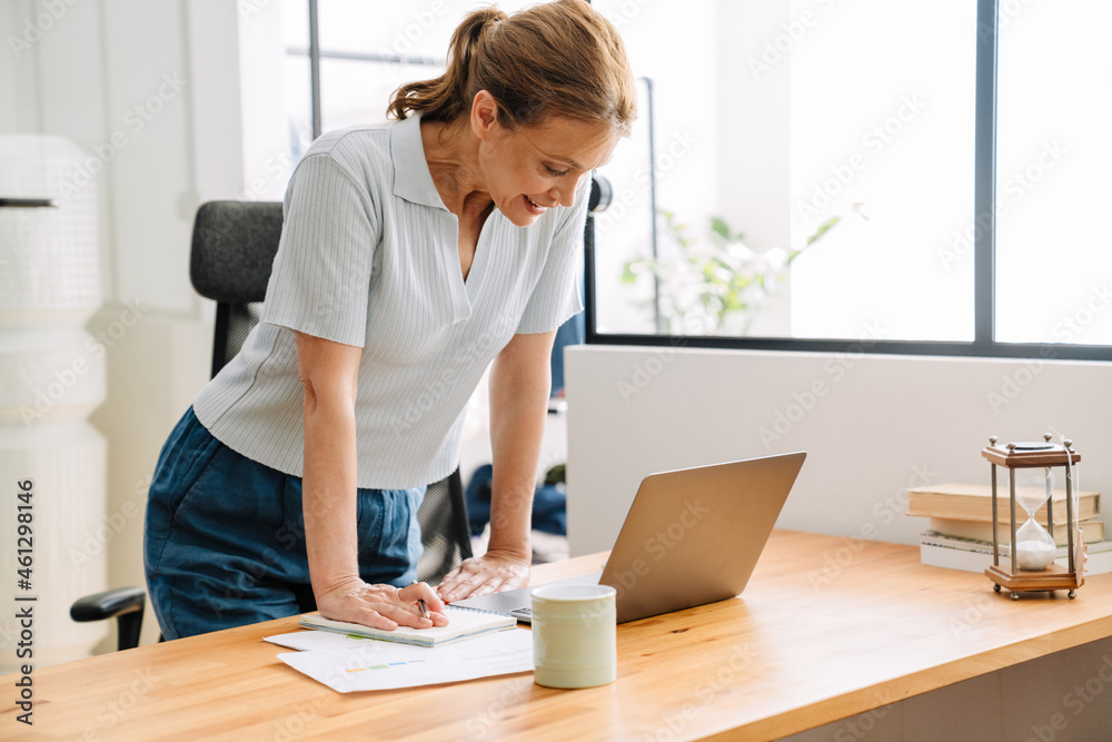 Ginger mature woman working with laptop while standing at desk Ginger mature woman working with laptop while standing at desk