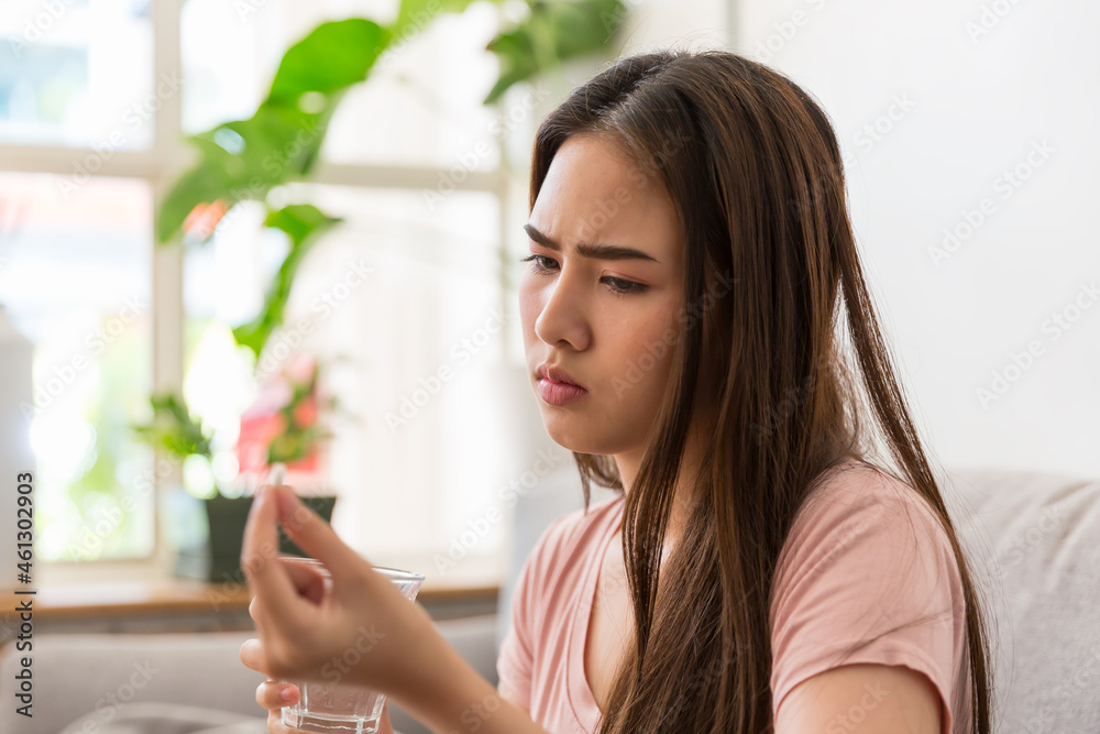 Young Asian woman feeling unwell and taking pill while sitting on sofa at home. Asian woman stressed and sick Young Asian woman feeling unwell and taking pill while sitting on sofa at home. Asian woman stressed and sick