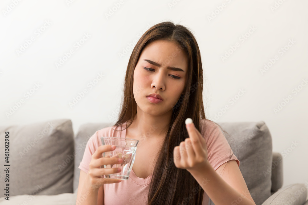 Young Asian woman feeling unwell and taking pill while sitting on sofa at home. Asian woman stressed and sick Young Asian woman feeling unwell and taking pill while sitting on sofa at home. Asian woman stressed and sick
