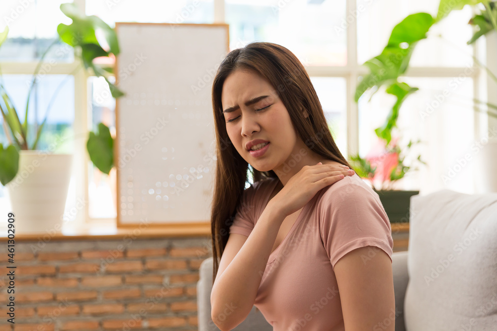 Young Asian woman feeling unwell and feeling pain her shoulder white sitting on sofa at home Young Asian woman feeling unwell and feeling pain her shoulder white sitting on sofa at home