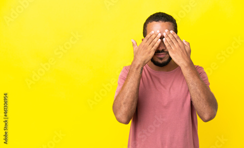 Young latin man isolated on yellow background covering eyes by hands