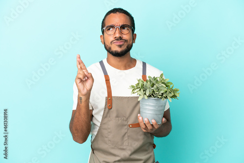 Gardener latin man holding a plant isolated on blue background with fingers crossing and wishing the best