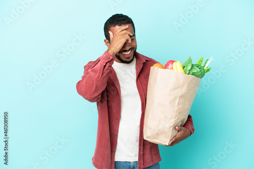 Young latin man holding a grocery shopping bag isolated on blue background laughing