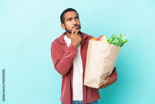 Young latin man holding a grocery shopping bag isolated on blue background and looking up